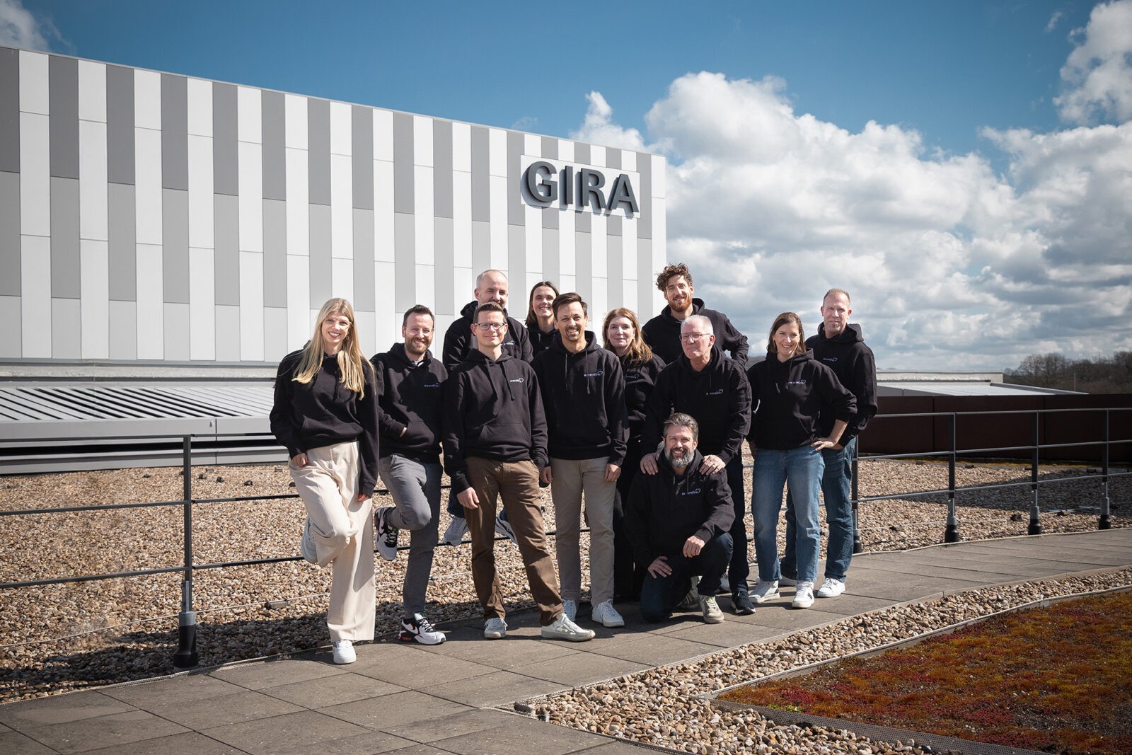 Gruppe von Menschen in schwarzen Hoodies steht draußen vor einem Gebäude mit dem Logo GIRA unter blauem Himmel.